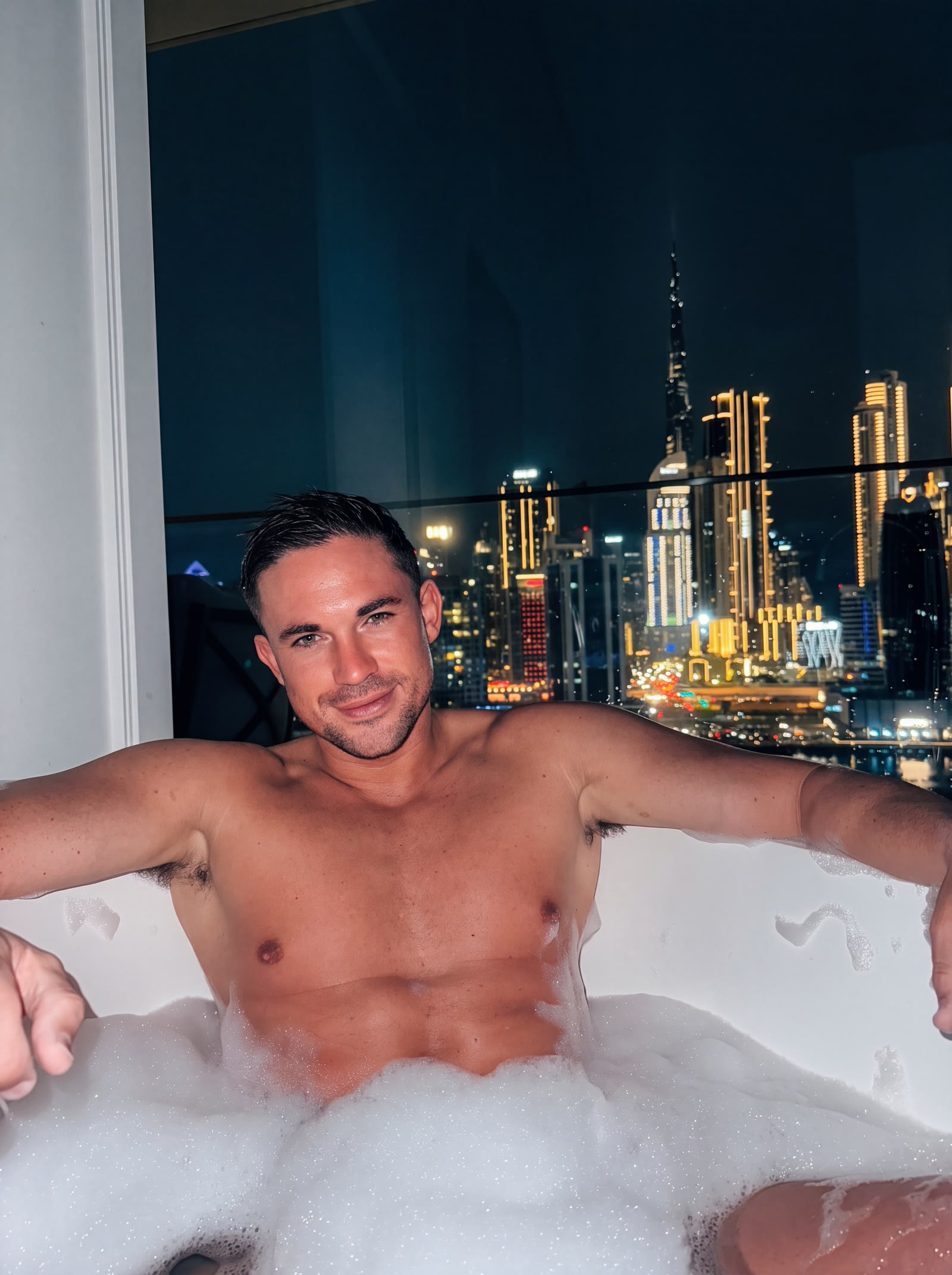 Will Parfitt in a bubble bath at night, a lit city skyline through the window behind him
